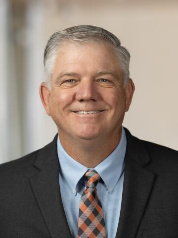 Photograph of a man wearing a black suit jacket, light blue dress shirt, and patterned tie with orange, black, and white colors. The background is softly blurred, focusing attention on the formal attire.