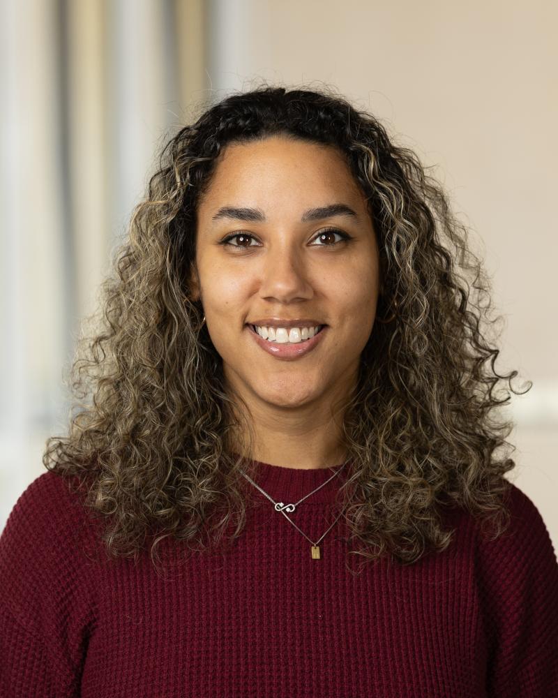 Photograph of a person with curly hair wearing a dark red sweater and two necklaces, one with an infinity symbol and the other with a small rectangular pendant. The background is softly blurred with neutral tones.