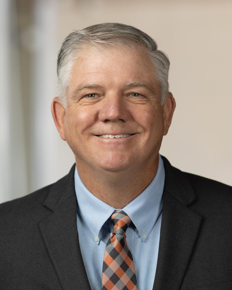Photograph of a man wearing a black suit jacket, light blue dress shirt, and patterned tie with orange, black, and white colors. The background is softly blurred, focusing attention on the formal attire.