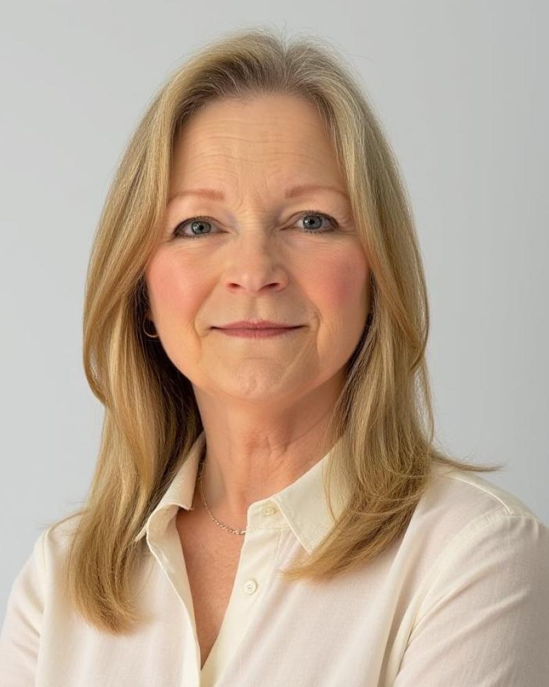 Photograph of a person with shoulder-length blonde hair wearing a cream-colored button-up shirt and a delicate necklace, set against a plain light gray background. 